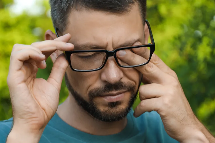 Allergic-Conjunctivitis-Clinic A man with a beard and glasses standing outside and wearing a blue t-shirt lifts his glasses and rubs his eyes due to allergy irritation. Get treatment for allergic conjunctivitis from Nonkulie Dladla, M.D. in Manhattan.