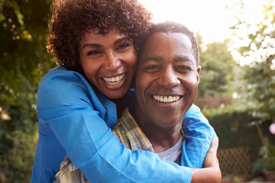 Autoimmune-Doctor A middle-aged African American couple outside, the woman embracing the man from behind pressing her cheek to his, both are smiling due to successful Autoimmune Disorder treatment from Nonkulie Dladla, M.D. in Manhattan.
