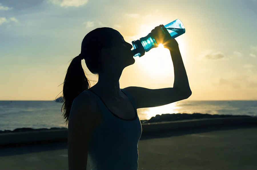 Chelation-and-Detoxification-Clinic A woman in exercise clothes drinking from a water bottle on the beach, with the water and sunset in the background. Get chelation and detoxification treatment from Nonkulie Dladla, M.D. in Manhattan.