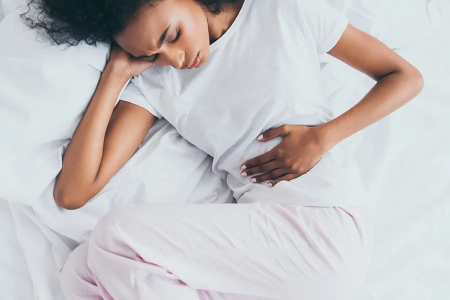 Crohns-Disease-Clinic A dark-skinned woman in white clothes lays on her bed clutching her middle before getting treatment for Crohn's Disease from Nonkulie Dladla, M.D. in Manhattan.