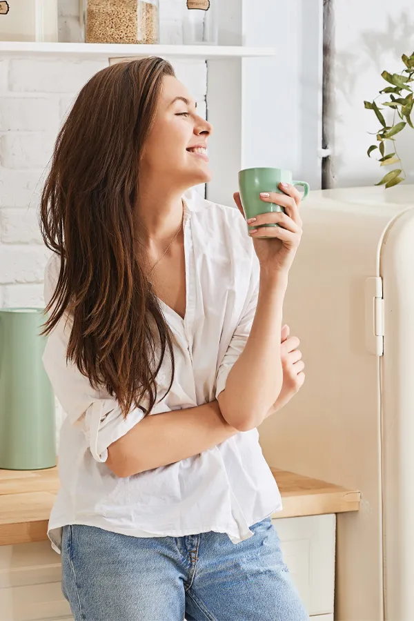 Crohns-Disease-Treatment A woman with a white blouse an jeans drinking tea in her kitchen, smiling after getting treatment for Crohn's Disease from Nonkulie Dladla, M.D. in Manhattan.