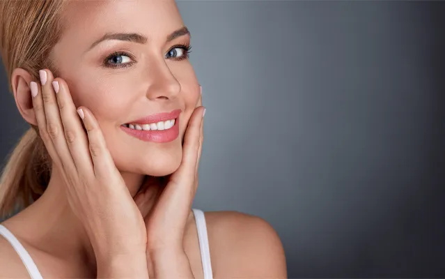 Facial-Clinic A middle-aged blonde woman with beautiful skin smiles and holds her face. Get facial treatments from Nonkulie Dladla, M.D. in Manhattan.
