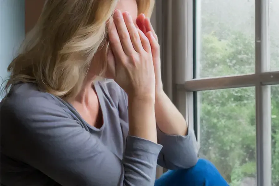 Graves-Disease-Clinic A blonde woman in a gray shirt sits in front of a window on a rainy day covering her eyes with her hands. Get treatment for Grave's Disease from Nonkulie Dladla, M.D. in Manhattan.