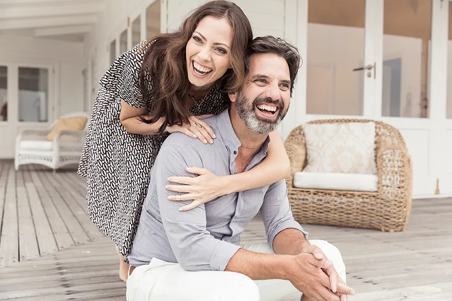 Health-and-Wellness-Services-Doctor A middle-aged couple on a wooden porch with wicker furniture; the woman is playfully hugging the man from behind. They are benefiting from the health and wellness services from Nonkulie Dladla, M.D. in Manhattan.