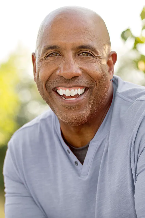 Healthy-Aging-Peptides-Treatment A middle-aged black man in a gray v-neck t-shirt sits outside smiling, happy with the healthy aging peptide treatments from Nonkulie Dladla, M.D. in Manhattan