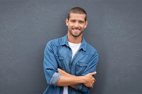 Hormones-for-Men-Doctor A man in a denim shirt stands smiling against a gray-blue wall, pleased with his testosterone hormone therapy from Nonkulie Dladla, M.D. in Manhattan.