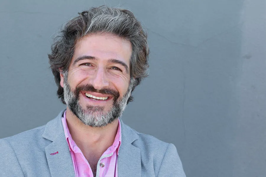 Hypogonadism-Clinic A smiling man with salt and pepper hair and beard wearing a pink shirt and gray blazer stands agains a gray wall. Get hypogonadism treatment from Nonkulie Dladla, M.D. in Manhattan.