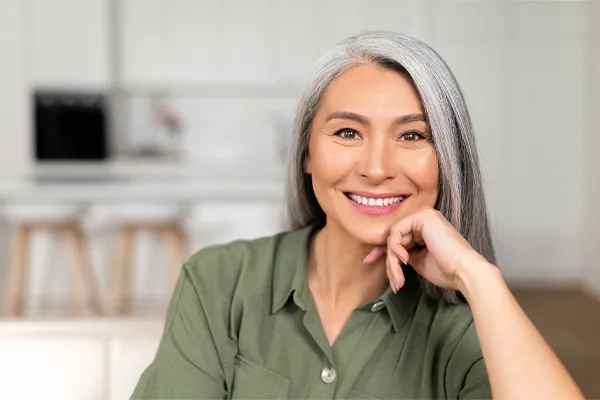 Menopause-Clinic An asian woman with long gray hair and a green shirt sitting and smiling on her couch, happy with her menopause care from [NAME} in Manhattan.