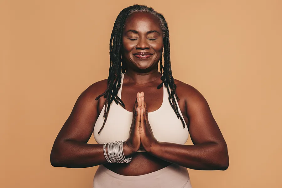 Obesity-Clinic An older African American woman confidently posing against a warm background smiling with hands in a prayer pose. Nonkulie Dladla, M.D. treats obesity using functional medicine in Manhattan.