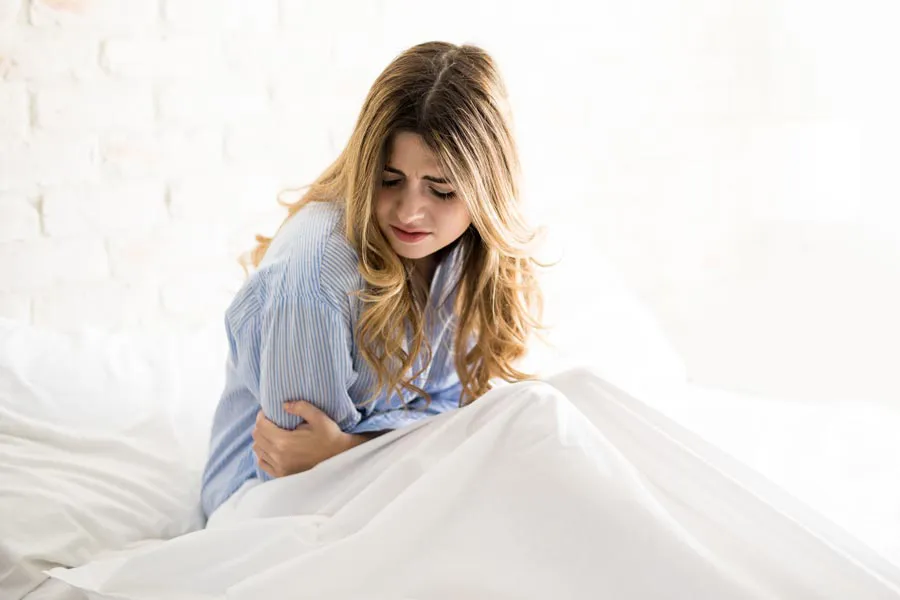 PMS-Therapy A woman with long blonde hair sits in her bed holding her stomach in pain, considering getting care for PMS from Nonkulie Dladla, M.D. in Manhattan.