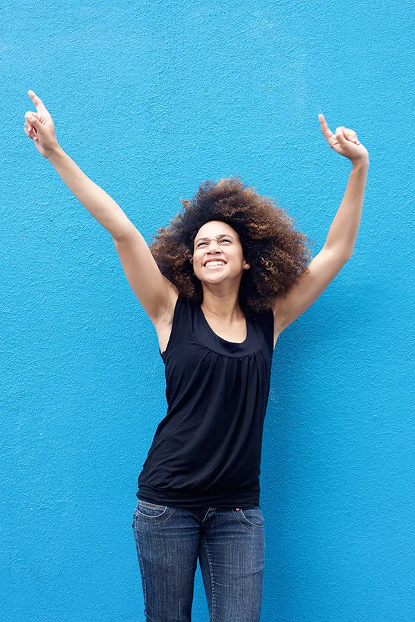 PMS-Treatment A woman in a dark blue tank top standing in front of a bright blue wall, raising her arms in celebration of relief from PMS from Nonkulie Dladla, M.D. in Manhattan.
