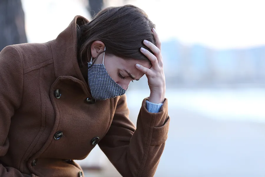 Post-Covid-Clinic A woman in a mask sits outside with her hand on her forehead in pain. Get long covid treatment from Nonkulie Dladla, M.D. in Manhattan.