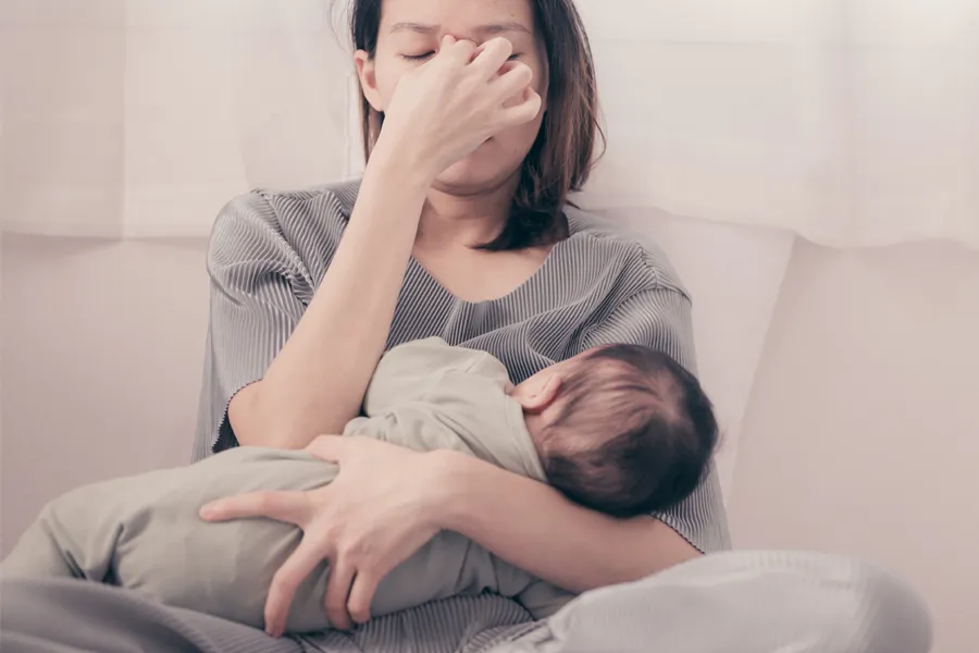 Postpartum-Hormone-Imbalance-Clinic A woman nursing an infant on couch rubs her eyes with exhaustion. Get treatment for postpartum hormone imbalance from Nonkulie Dladla, M.D. in Manhattan.