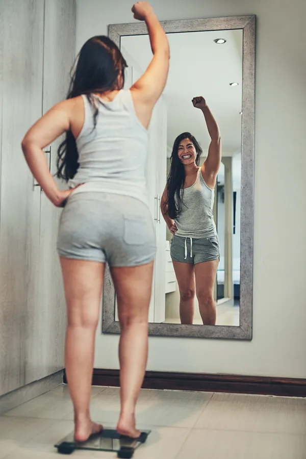 Tirzepatide-Treatment A woman in workout clothes stands on a scale looking in the mirror and pumping her hands in celebration of her successful Tirzepatide treatment from Nonkulie Dladla, M.D. in Manhattan.